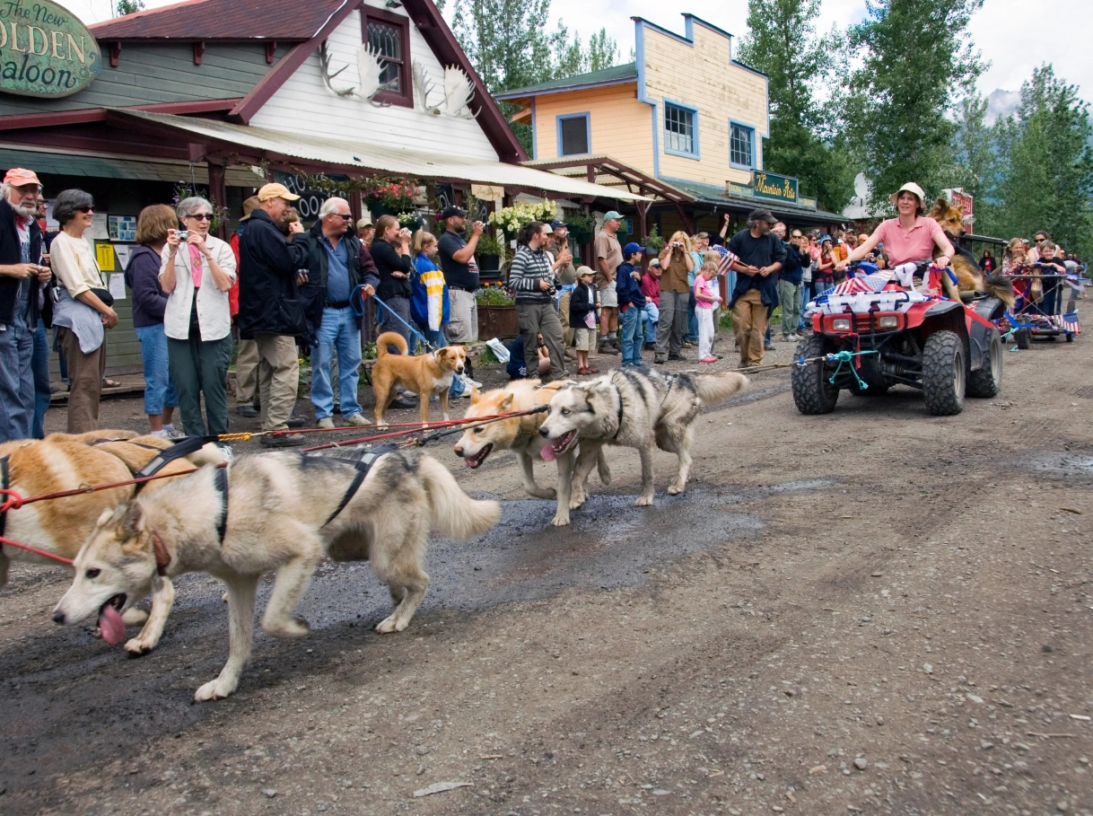 Sled dogs pulling an ATV in a parade on a dirt road with spectators watching