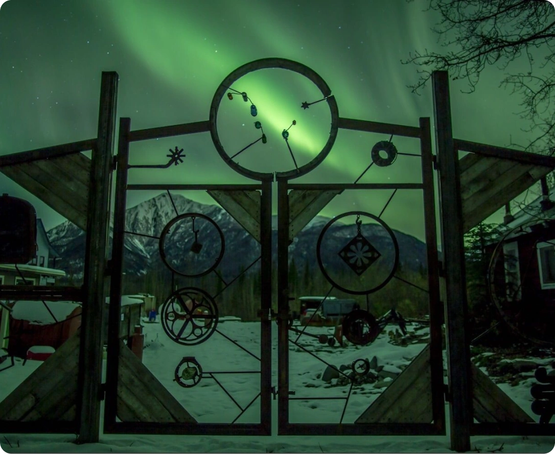 Aurora borealis behind a decorative gate with circular metal ornaments, in a snowy mountainous landscape