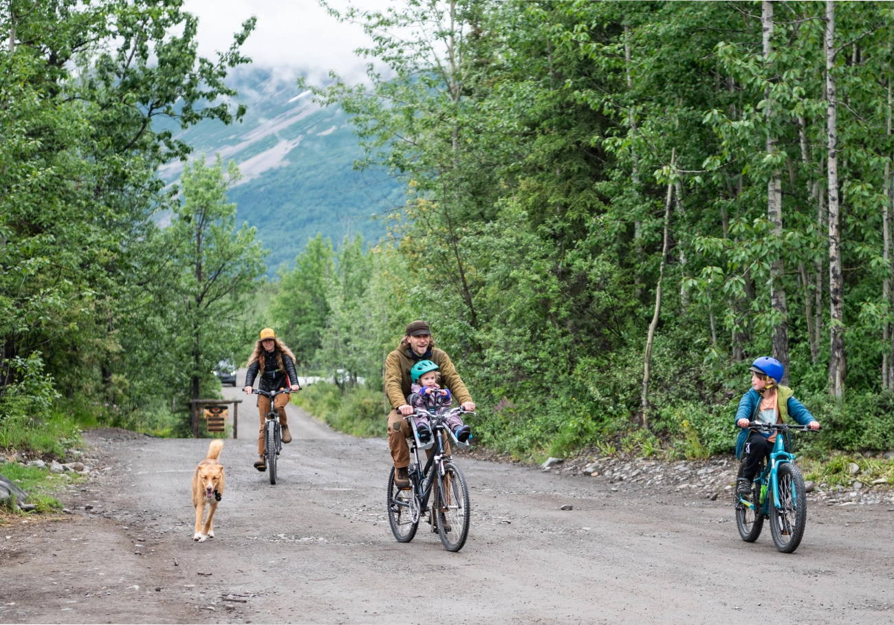 Family biking on a forest trail with a dog, surrounded by lush green trees and a mountain backdrop