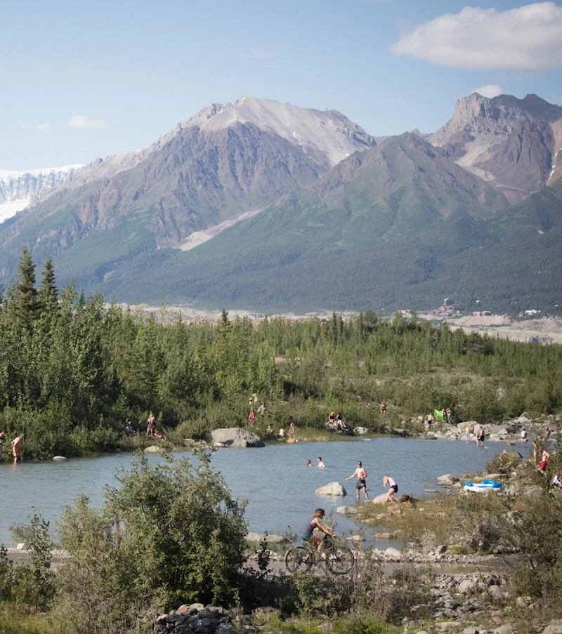 People enjoying outdoor activities by a river with a backdrop of mountains