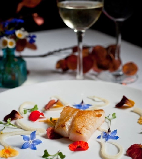 Gourmet dish with seared fish and edible flowers, wine glasses in backdrop