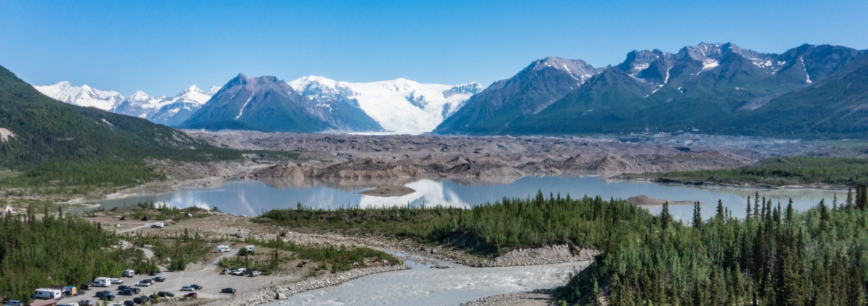 Scenic view of a glacier, lake, and a campground with vehicles and tents in the foreground